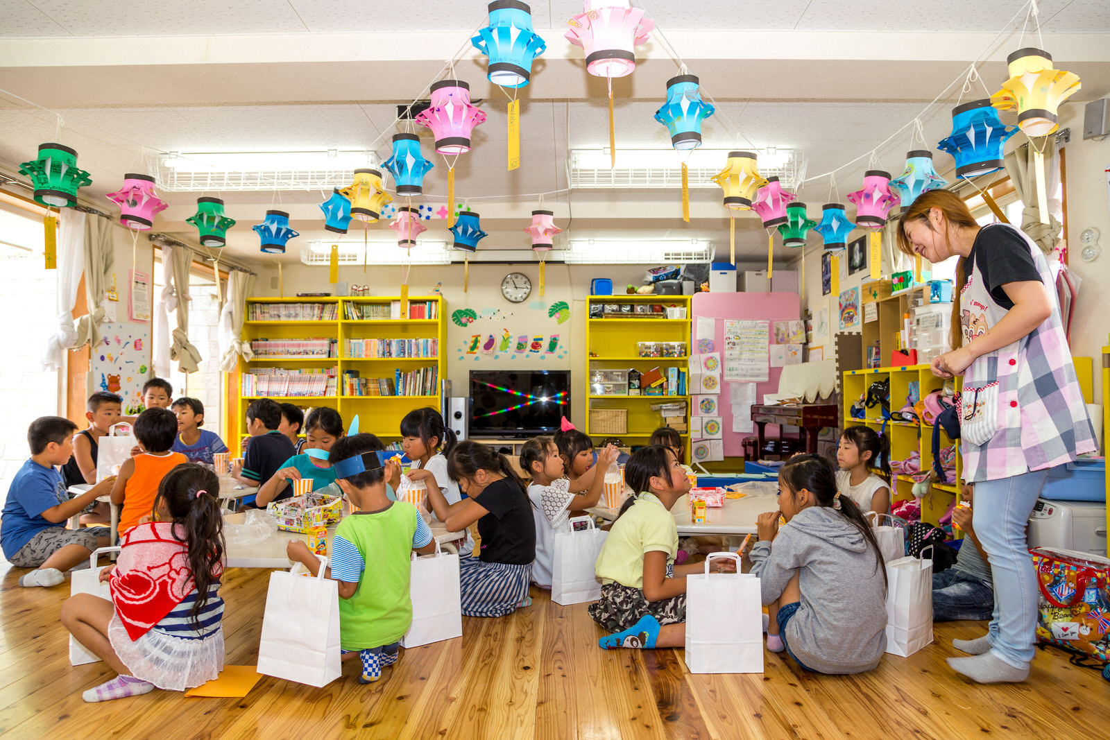 色とりどりの色画用紙で作られた提灯が天井から吊るされた明るい室内で、たくさんの子どもたちが机を囲んでお菓子を食べたり会話を楽しんでいる写真