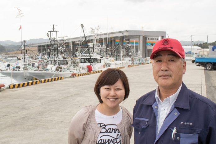 数隻の漁船が停泊している港に、リポーターの女性と佐藤さんが並んで立っている写真