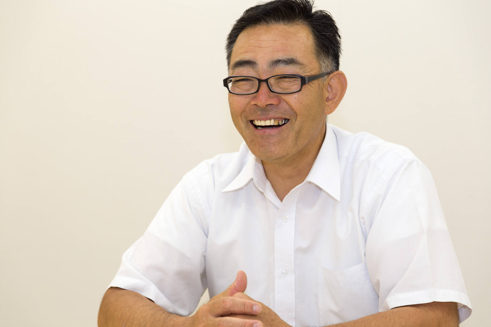 テーブルに両手を組み笑顔で座っている白い半袖シャツを着た高橋さんの写真