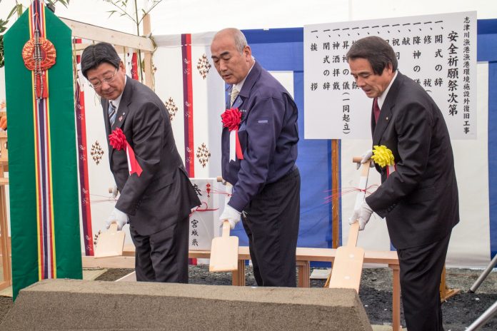 スーツ姿の男性二人と作業着の上着を着た男性が、白手袋をはめて儀式用の木のスコップで砂山に鍬入れをしている安全祈願祭の写真