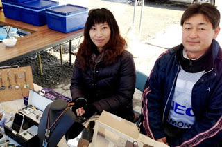 屋外のテーブルの上にネックレスやイヤリングなどのジュエリーが丁寧に並べられ、その後ろに防寒着を着た女性とNECの白いビブスを着た男性が並んで座っている写真