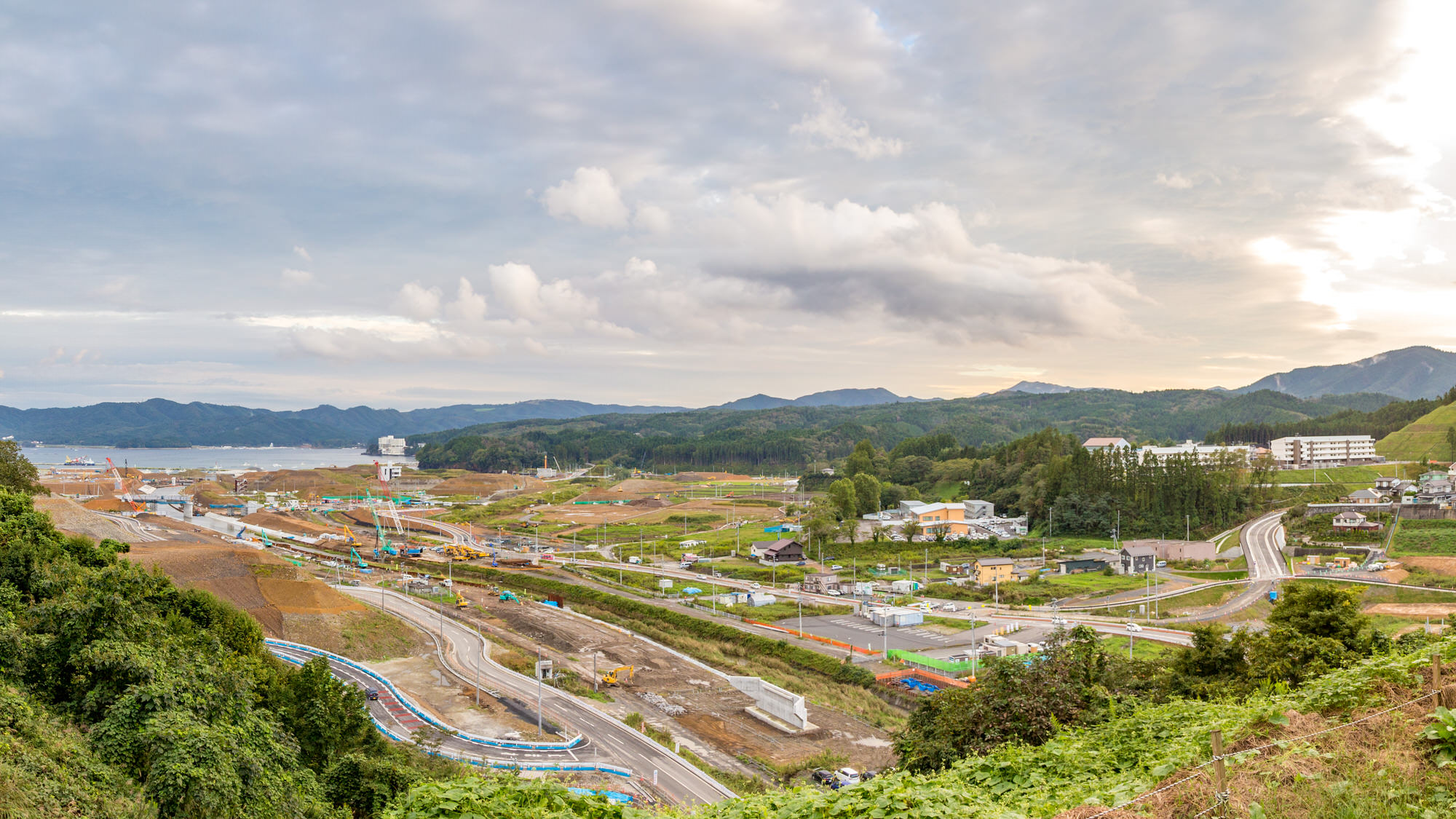 手前には緑に覆われた丘陵地帯が広がり、その奥には道路や点在する建物が見え中央には工事中のエリアがあり、重機や資材が配置されている広々とした田園風景と建設現場が一体となった様子を写した写真