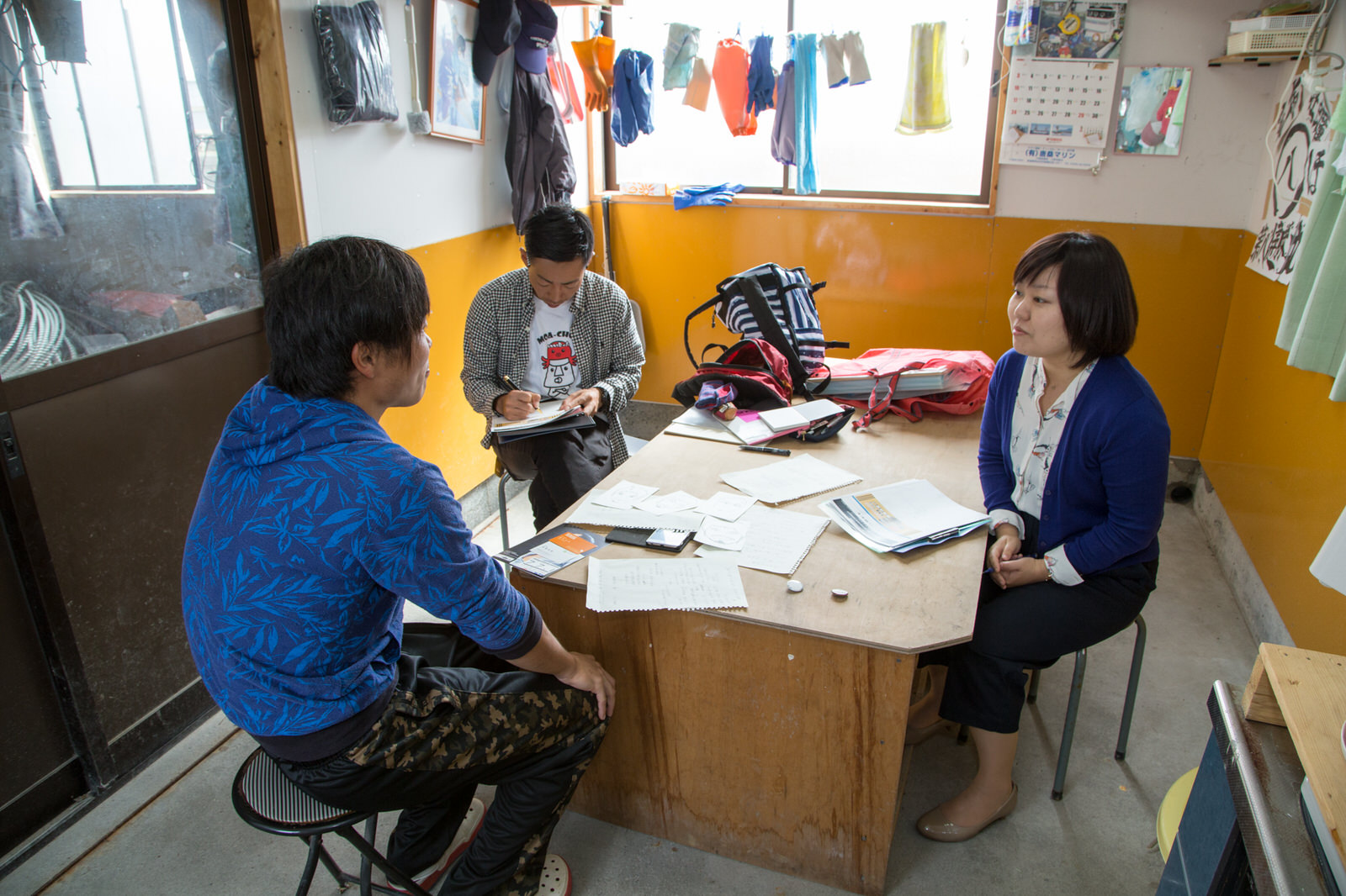 窓辺にゴム手袋やタオルなどがたくさんかけられた作業スペースの木の台を囲んで、浅野さんと女性リポーター、メモを取っている男性が取材を行っている様子の写真