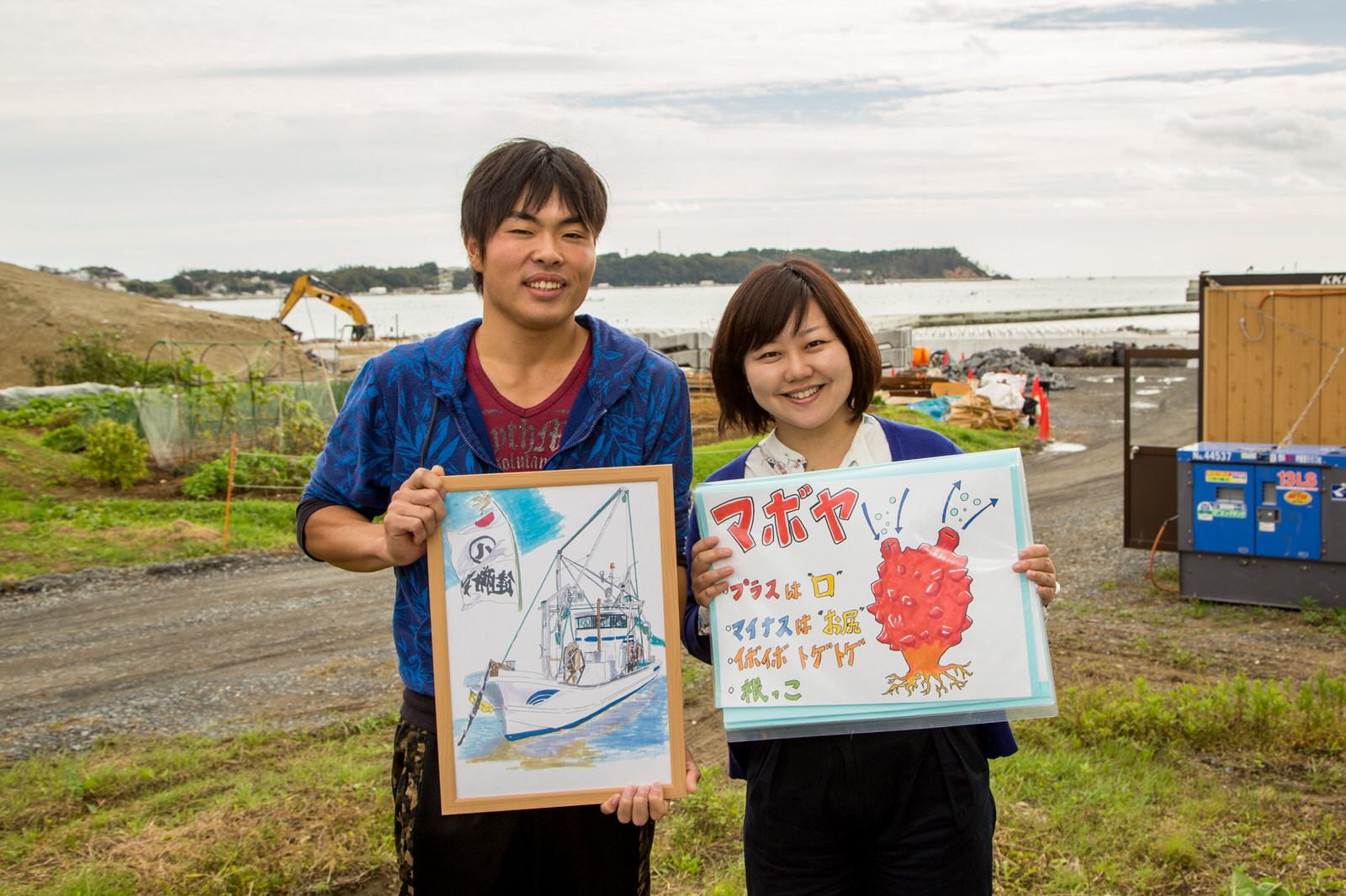 工事中の海岸を背景に漁船の絵を持った浅野さんと、マボヤのイラストが描かれた紙を持った女性リポーターが並んで立っている写真