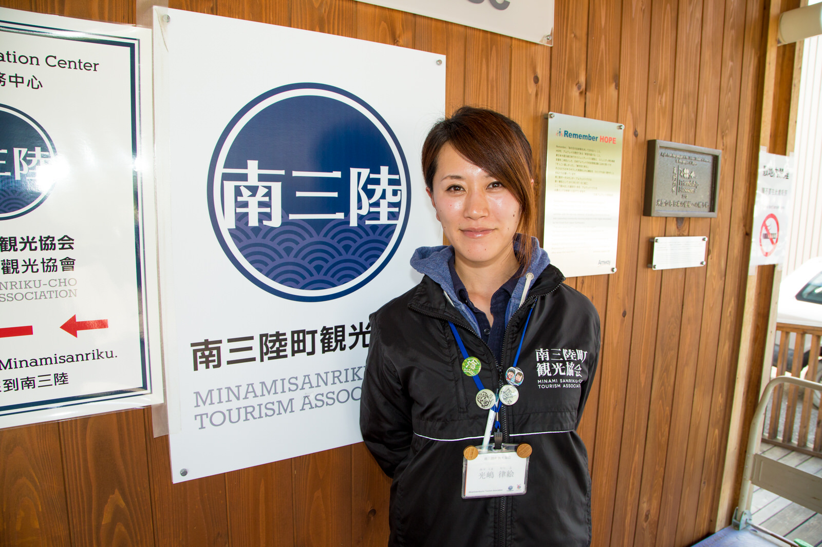 南三陸観光協会の看板が設置された壁の前に笑顔で立っている光嶋さんの写真