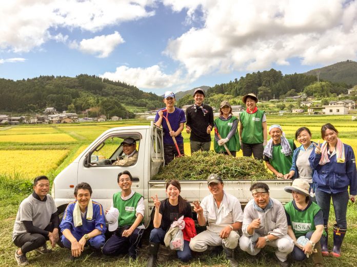 山々が広がっている黄金色の田んぼの前で、トラックの荷台に積まれた草の前で農作業を終えた男女15人が笑顔でポーズをとり並んでいる写真