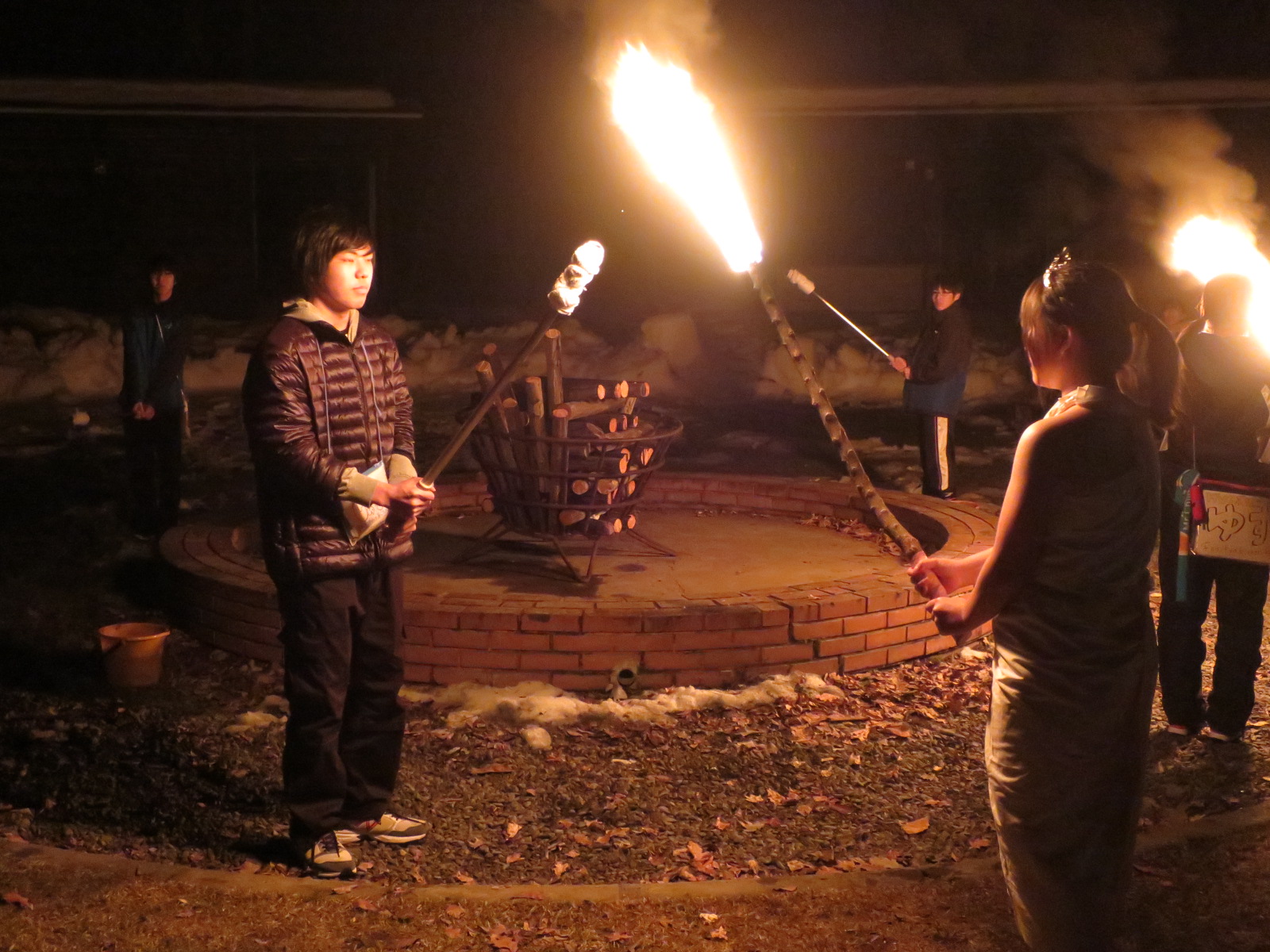 夜の屋外で複数の人々が薪の束が組まれた焚き火台を囲みながら、長い棒の先に火のついた松明を持って立っている様子を写した写真