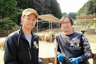 羊のいる柵の前で、2人の男性が並んでカメラを見つめている様子をアップで撮影した写真