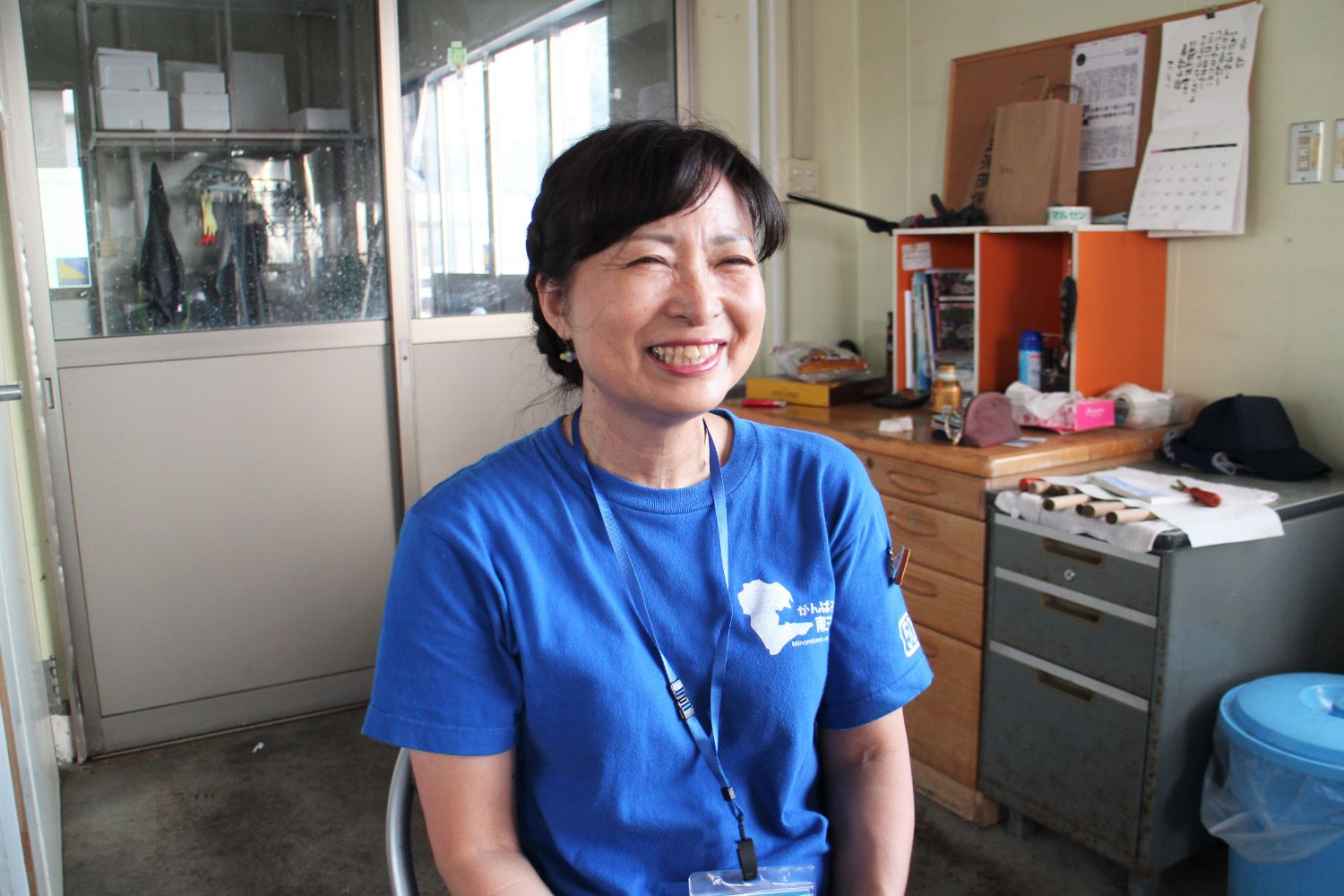 室内の事務所のような場所で、イスに座った笑顔の青いTシャツを着た女性をアップで撮影した写真