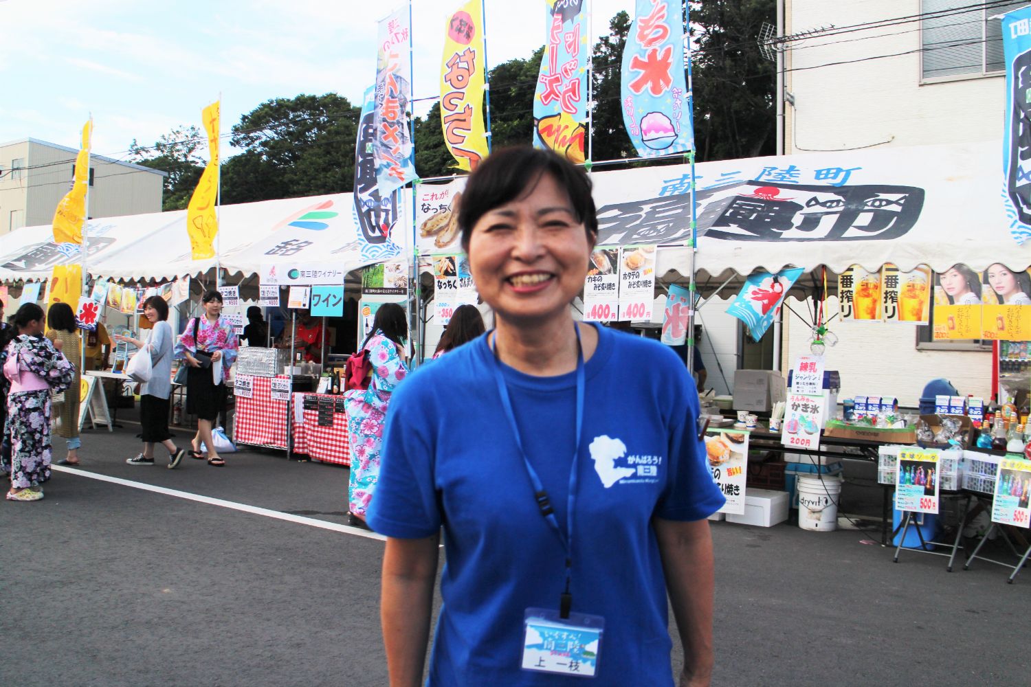 カラフルなのぼり旗や浴衣姿の来場者、飲食の屋台などが並ぶお祭り会場で、青いTシャツを着た女性がでカメラに向かって笑顔で立っている写真