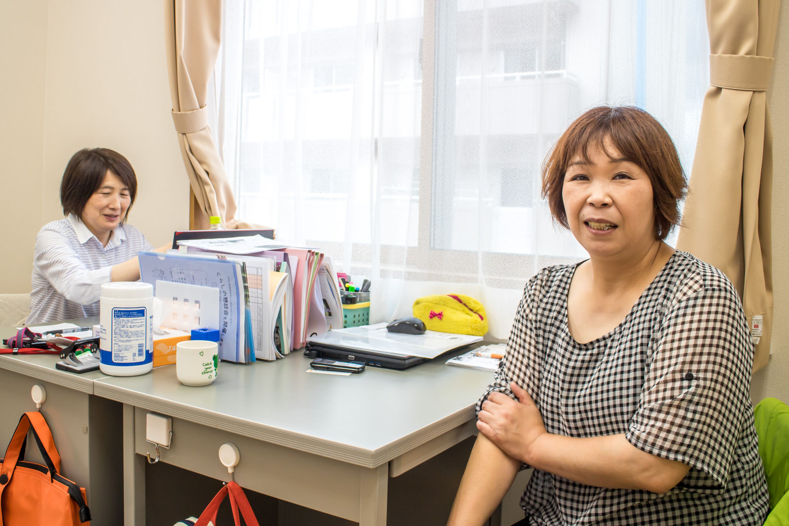窓際に置かれた対面の事務机の中央にたくさんの書類が立てられ、席に着いた2人のうち奥の女性が書類に手を伸ばし、手前の女性はカメラに笑顔を見せている写真