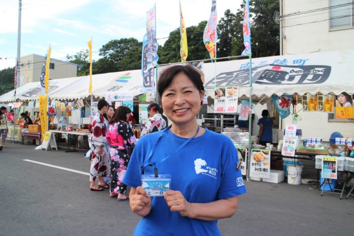 屋台が並ぶお祭り会場で、青いTシャツを着た女性が名札を見せながら笑顔で立っている写真