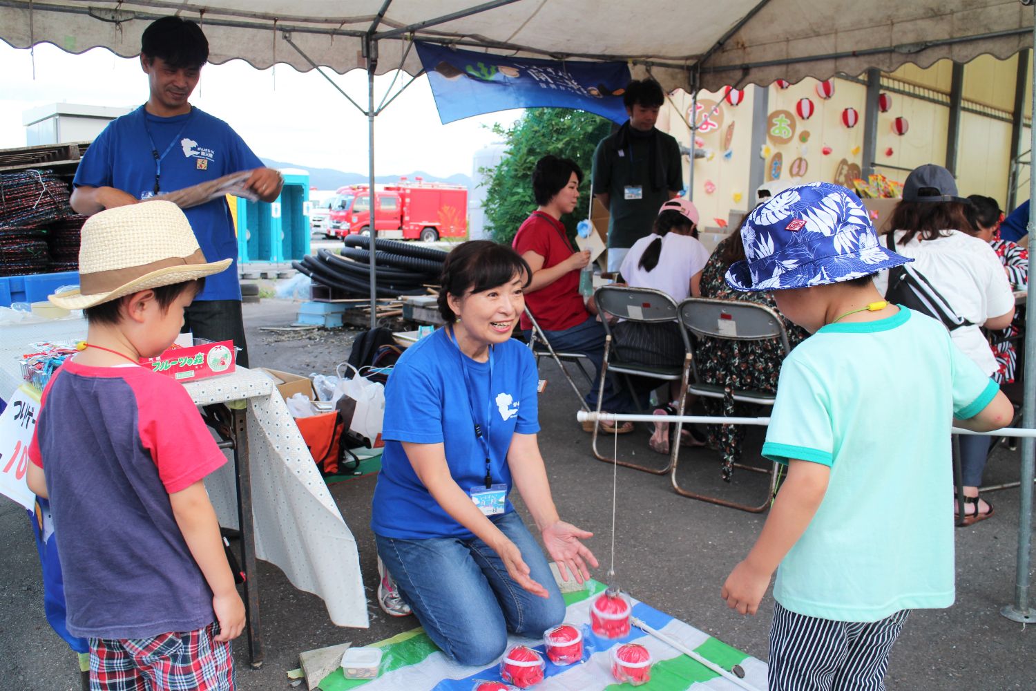 青いTシャツを着た女性がテントの下で2人の子どもと一緒におもちゃの釣りゲームをしており、楽しそうにしゃがんで子どもたちに話しかけている様子が写っている写真