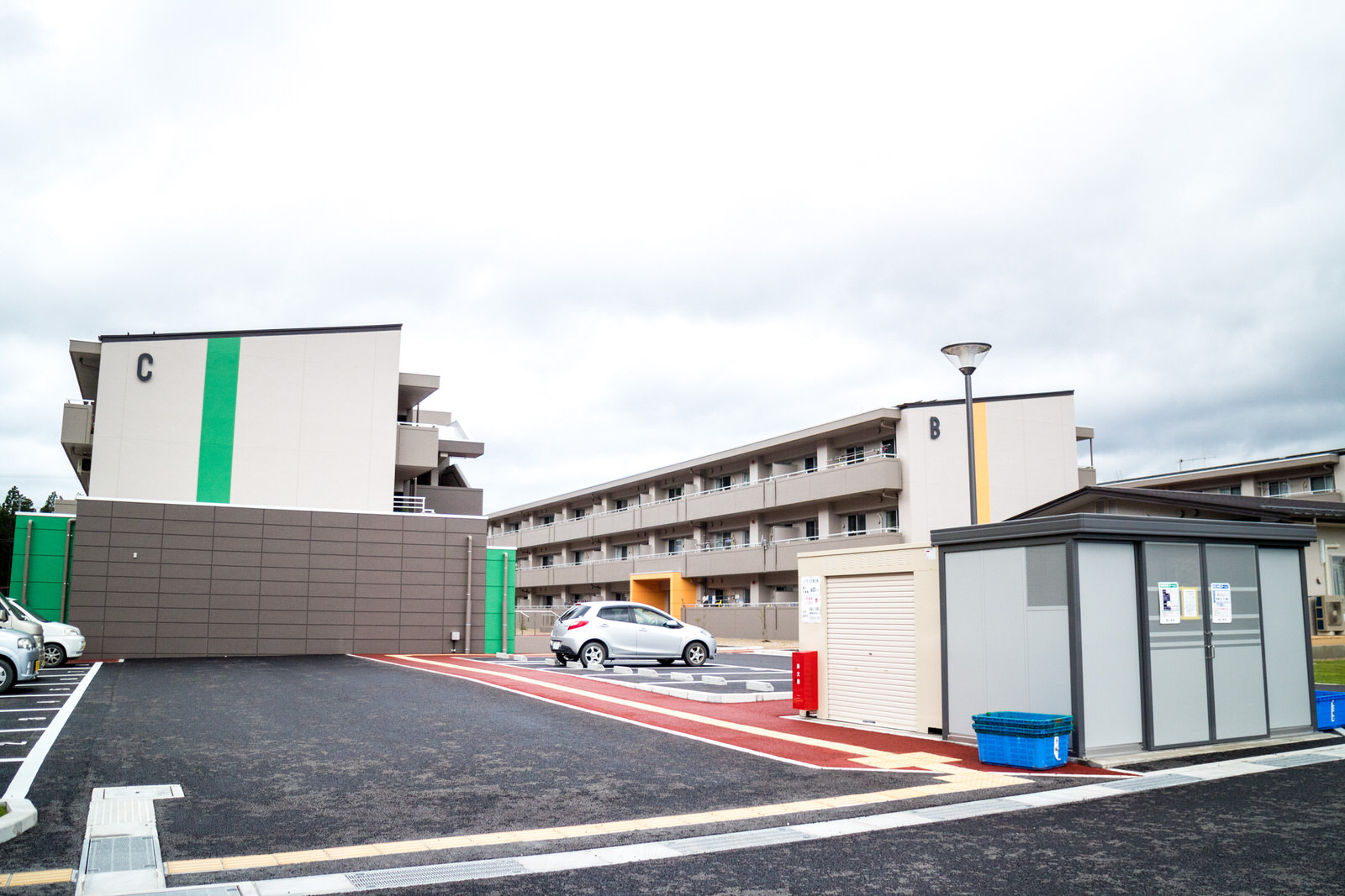 広い駐車場の一角に倉庫のような建物があり、駐車時用の先には白い建物に緑色の縦ラインが入った建物と黄色の縦ラインが入った建物が2棟並んでいるのが見える3階建ての災害公営戸倉住宅の写真