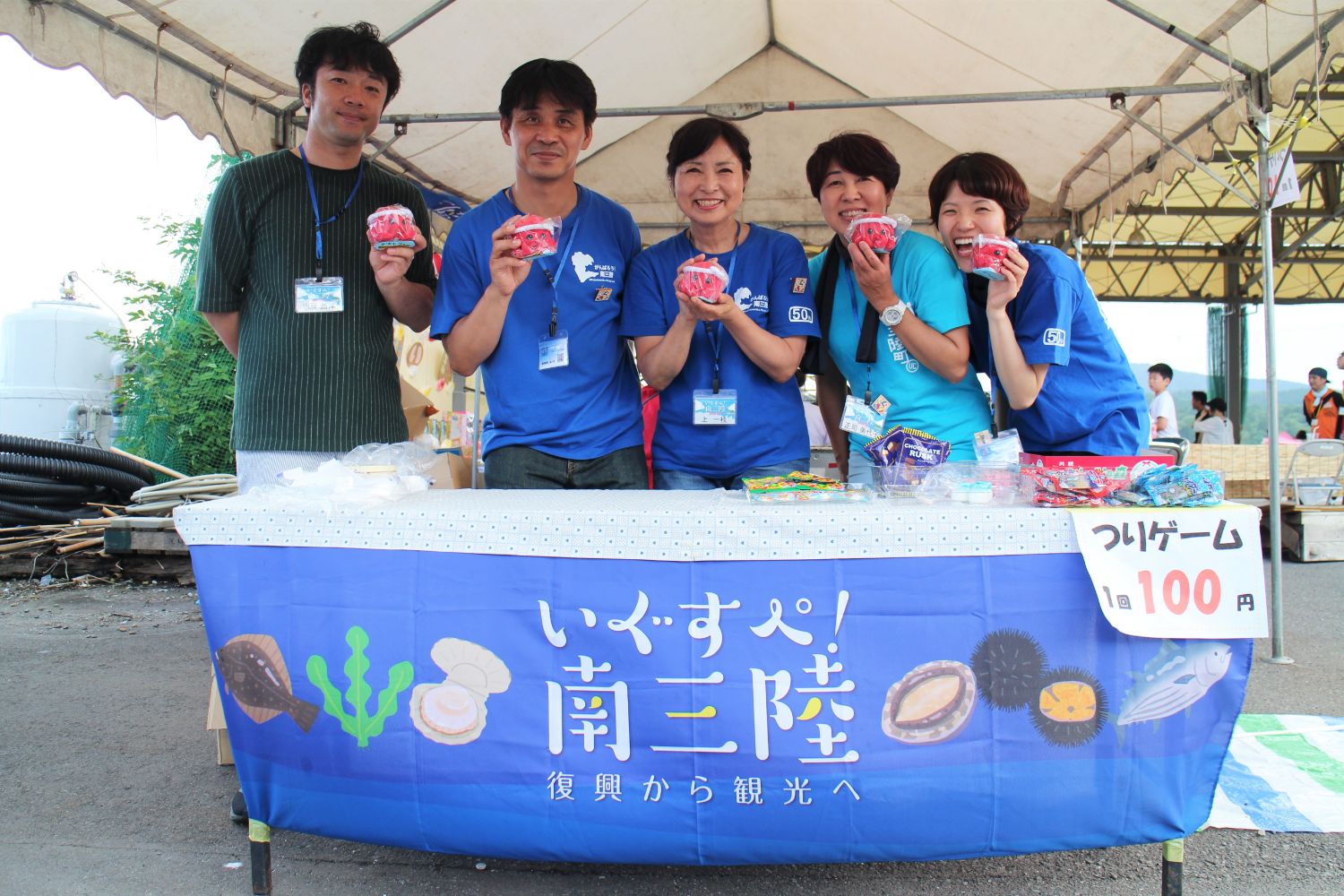 「いぐすぺ！南三陸 復興から観光へ」と書かれた青いテーブルカバーがかかった釣りゲームのブースで、手に赤いオクトパス君のぬいぐるみを持った青いTシャツを着た5人のスタッフが笑顔で横一列に並んでいる写真