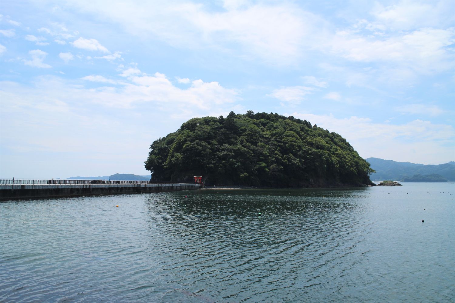 海に突き出た防波堤の先に、緑豊かな小さな島があり、その入口付近には赤い鳥居が建っている写真