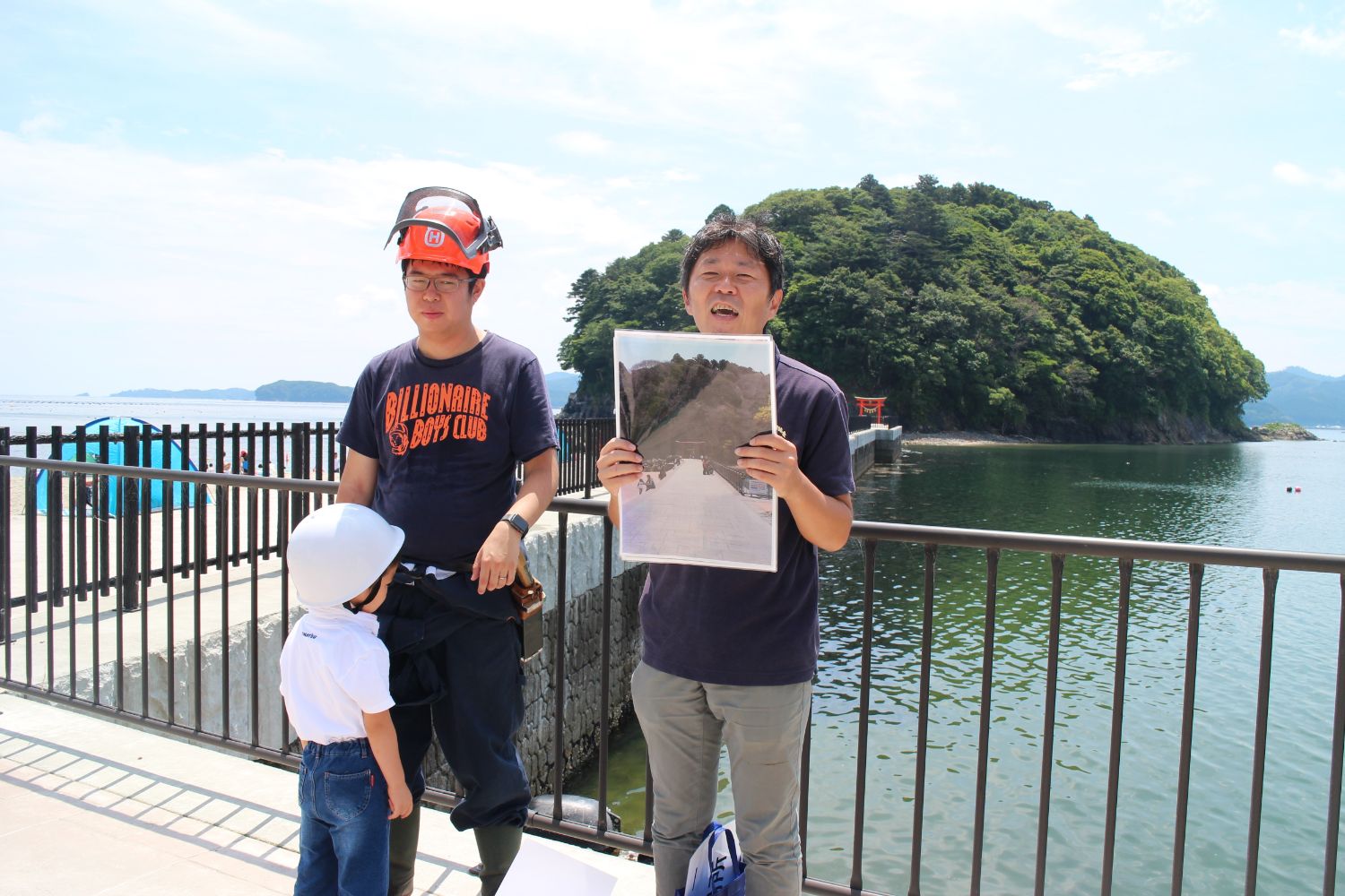 海辺の遊歩道で、白いヘルメットを被った男の子が紺色のTシャツを着た佐藤さんに話しかけており、その隣では別の男性が橋と鳥居が写った大きな写真を手に持って説明している様子の写真