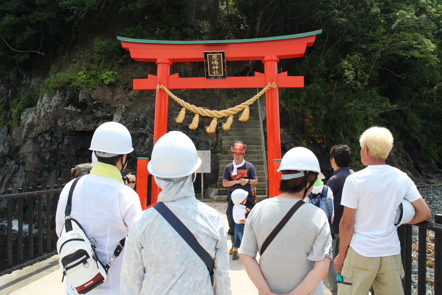 赤い鳥居の前に立つ案内役の佐藤さんがヘルメットを被り、数名のヘルメットを着用した参加者たちに向けて説明をしており、鳥居の奥には石段が続き、背後の岩肌と木々に囲まれた神聖な雰囲気の中で見学が行われている写真