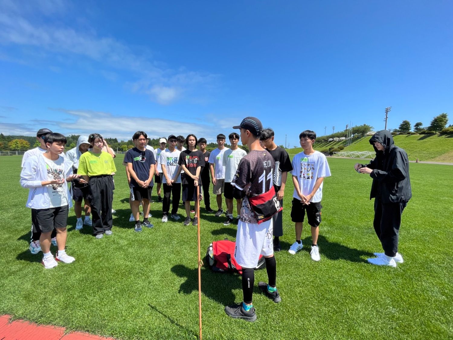 青空の下、芝生のグラウンドに集まった参加者たちが円形に立ち、中央で説明をしている人物の話を真剣に聞いている様子の写真