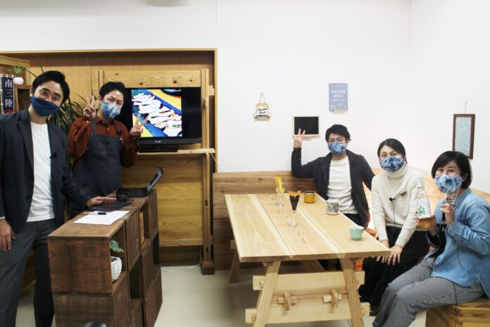 木製のテーブルに3人が座り、カウンターには2人が立っていてカメラ目線で笑顔で写っている写真