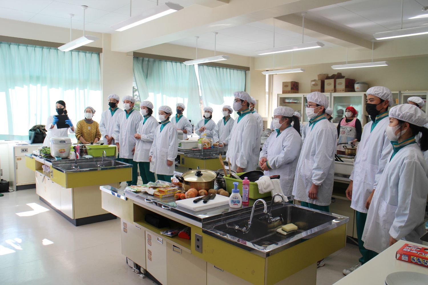 調理室でたくさんの生徒たちが白衣とマスク、帽子を身に着け、調理器具や食材が並べられている調理台の前に並んで前を向き、話を聞いている様子の写真