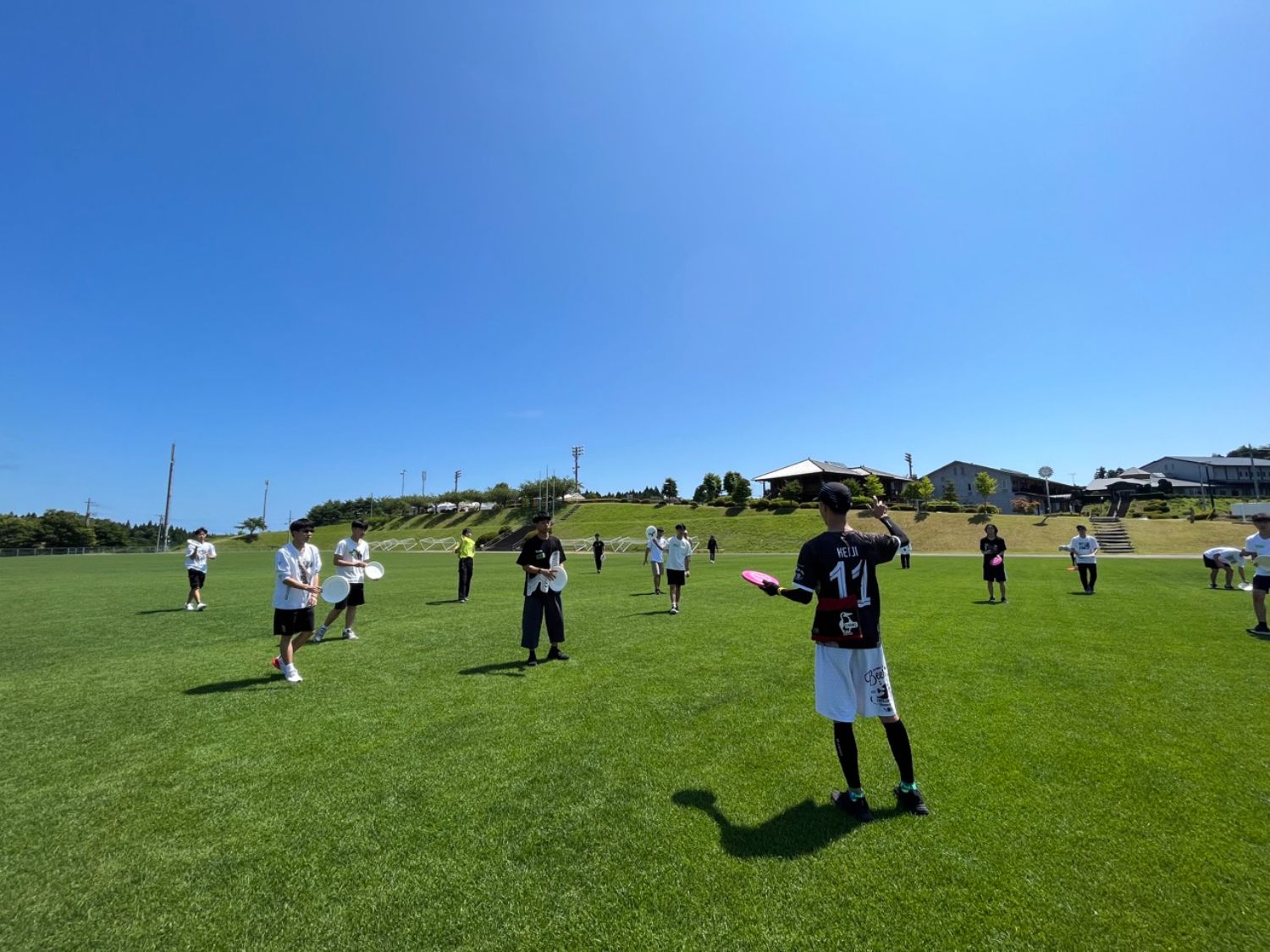 晴天の屋外の芝生のフィールドで、多くの若者がフリスビーを持ちながら前方の男性の説明を聞いている様子の写真