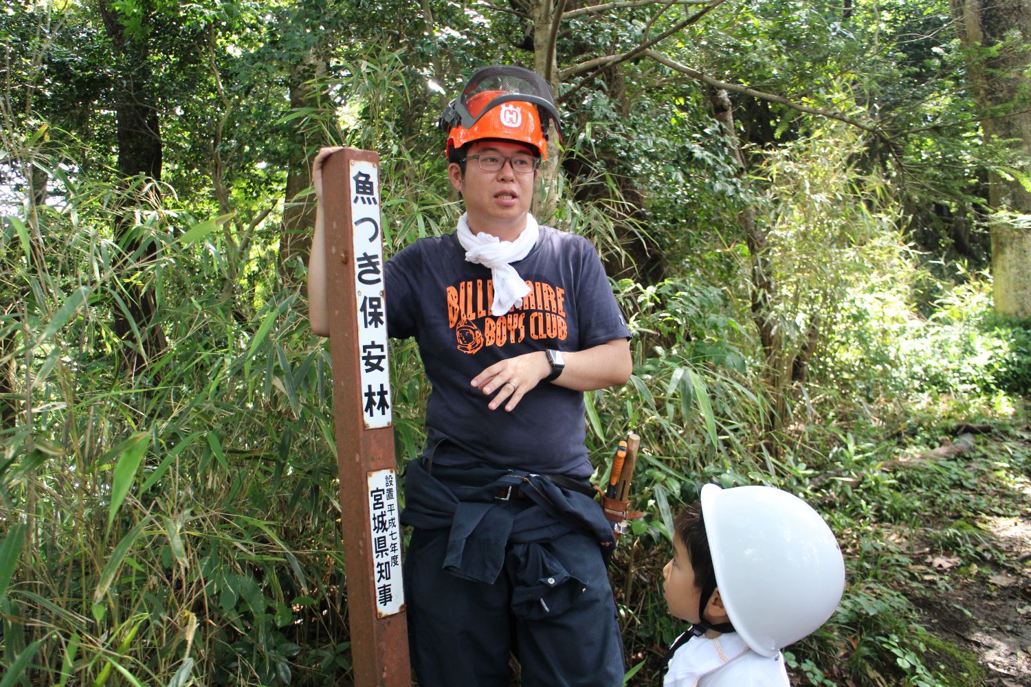 森の中で「魚つき保安林」と書かれた木製の標柱の横に、オレンジ色のヘルメットを被った佐藤さんが立ち、足元では白いヘルメットを被った子どもが見上げて話を聞いている写真