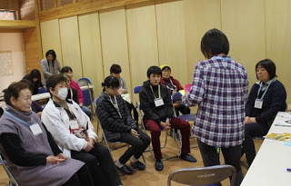 青いチェック柄のシャツを着た女性が、椅子に座った参加者たちの方を向いて立っている写真