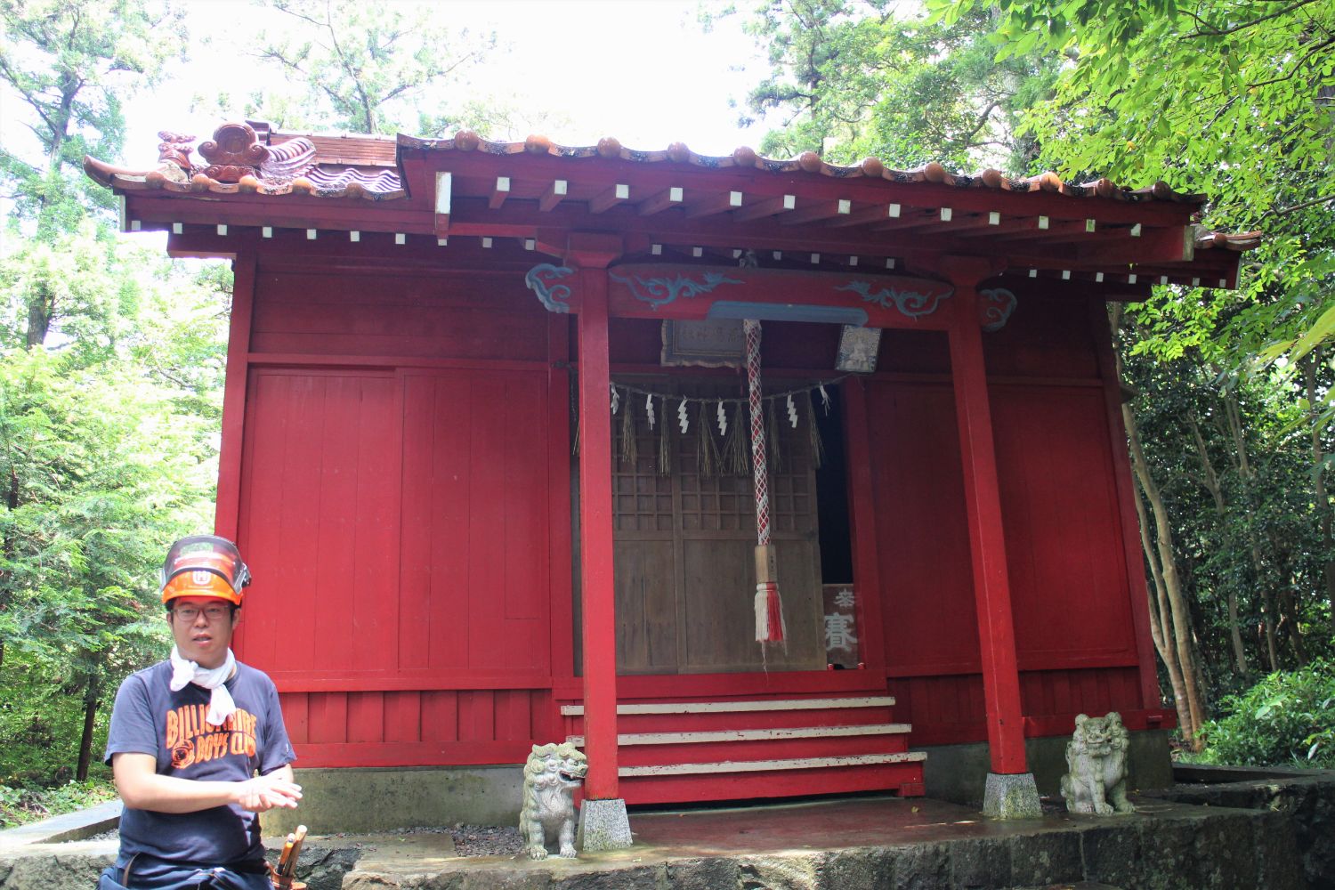 森の中にある正面にしめ縄と鈴が垂れ下がり、左右に狛犬が配置されている朱色の小さな神社の前で、オレンジ色のヘルメットを身につけた佐藤さんが手を動かしながら説明している写真