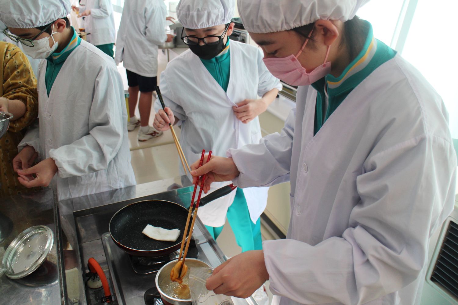 白衣と帽子、マスクを着けた生徒が、フライパンと鍋が置かれた調理台で作業をしており、1人の生徒が鍋でゆでたホヤを長い菜箸ですくっている調理作業中の写真