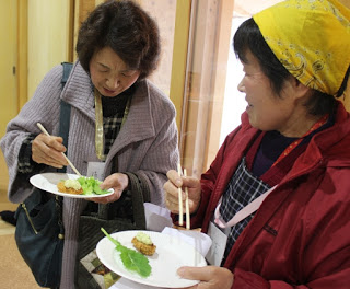 生の葉野菜と揚げ物が乗った紙皿を手に、笑顔で試食をしている女性2人の写真