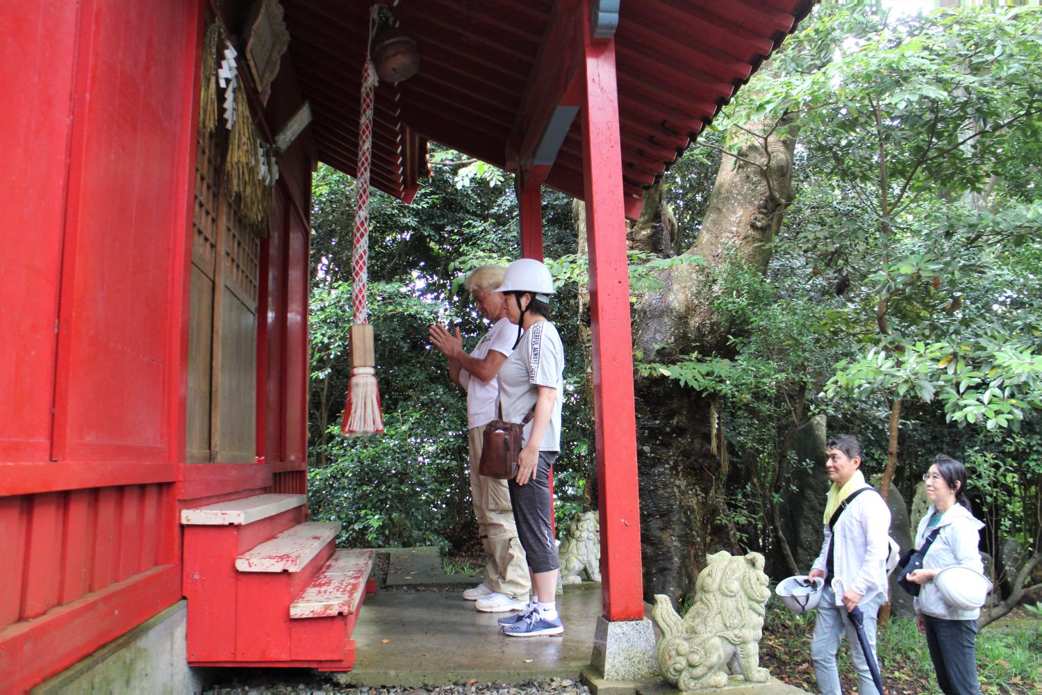まわりを緑に囲まれ、足元には狛犬が座っている赤い小さな神社の前でヘルメットを被った男女が手を合わせて参拝し、その後ろに順番を待つ人々が並んでいる写真