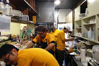 厨房内で黄色いシャツを着た複数のスタッフが料理の仕込みをしている様子の写真