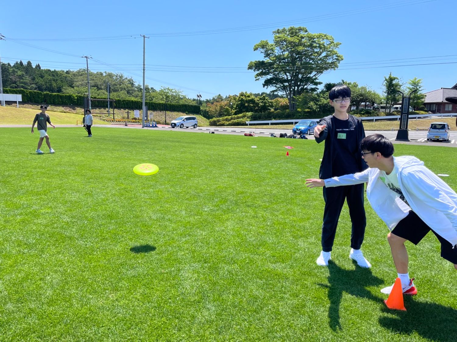 芝生の広場で、白い上着を着た若者が前かがみの姿勢でフリスビーを投げ、黒いシャツを着た学生が指導している様子を写した写真