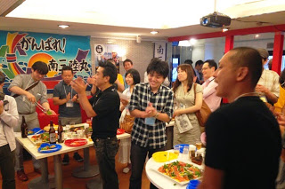 テーブルの上に料理やビール瓶が置かれた室内で多くの人が集まり、拍手しながらイベントを楽しんでいる様子の写真