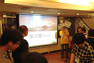 プロジェクターで「南三陸Now」と表示されている前で、黄色のシャツを着た男性スタッフがマイクを持って進行している様子の写真