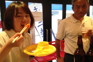 女性が笑顔で箸を使って料理を食べ、隣にはビールが注がれた透明のグラスを持った男性が立ち食事を楽しんでいる様子の写真