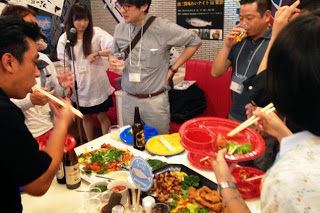 テーブルの上に料理が並び、参加者が箸で料理を取りながらビールを飲み楽しんでいる様子の写真