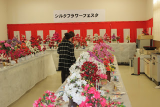 たくさんの繭細工の花が展示された長机の間の通路に立った2人の女性が作品に見入っている写真