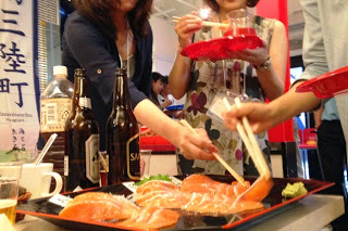 数人の参加者が赤い皿を持ちながら、箸でサーモンの刺身をつまんで試食している様子の写真