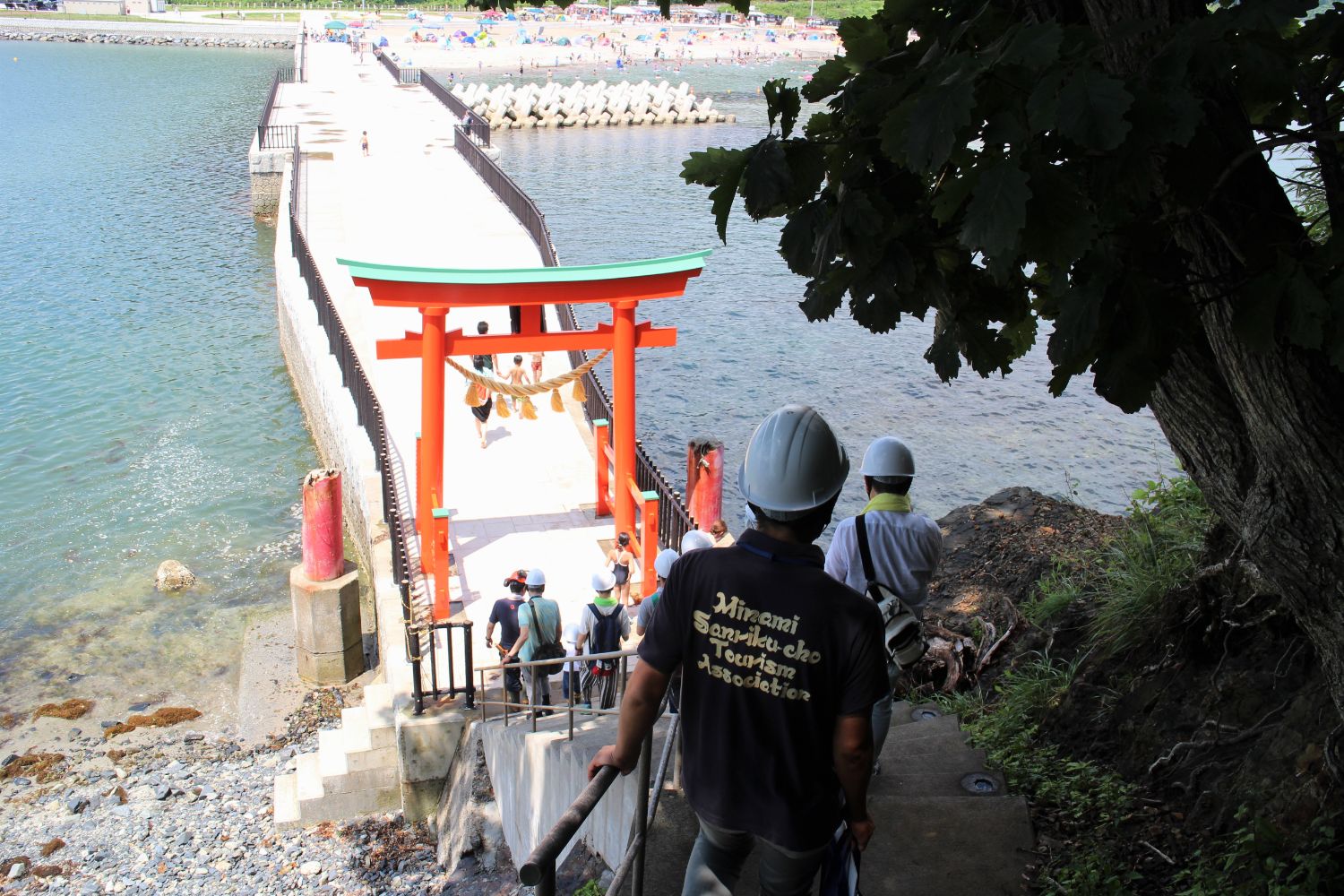 ヘルメットを被った参加者たちが石段を降りながら赤い鳥居をくぐり、海へと続く桟橋に向かって歩いている様子を後方から撮影した写真