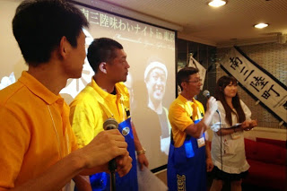 スクリーンを背景に、オレンジ色のシャツの男性と、白色の服を着た女性の間に立つ黄色いシャツと青いエプロンを着た2人の男性の内、右側の男性がマイクを持って話している様子の写真