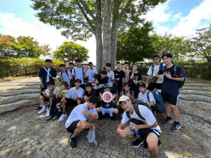 広場の大きな木の下で、リュックを背負った若者たちが並んで座ったり立ったりしながら、笑顔でカメラに向かってポーズを取っている集合写真