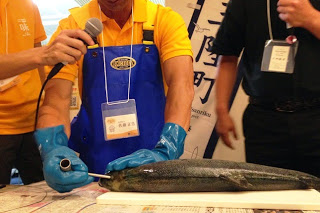 青い手袋とエプロン姿の男性が魚に長い針金を当てて、神経抜きの実演をしている様子の写真