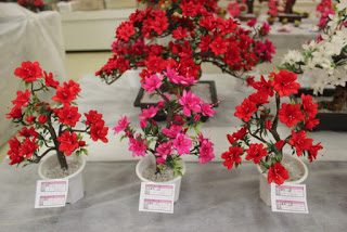 手前に赤とピンクの花の小ぶりな繭細工が3つ並び、奥には赤や白の大きめの繭細工の花が展示されている写真