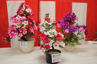 紅白幕の前に、薄いピンクから濃いピンクのグラデーション、赤と白、ピンクと紫の違う色を組み合わせた繭細工の花の作品が3つ並んでいる写真