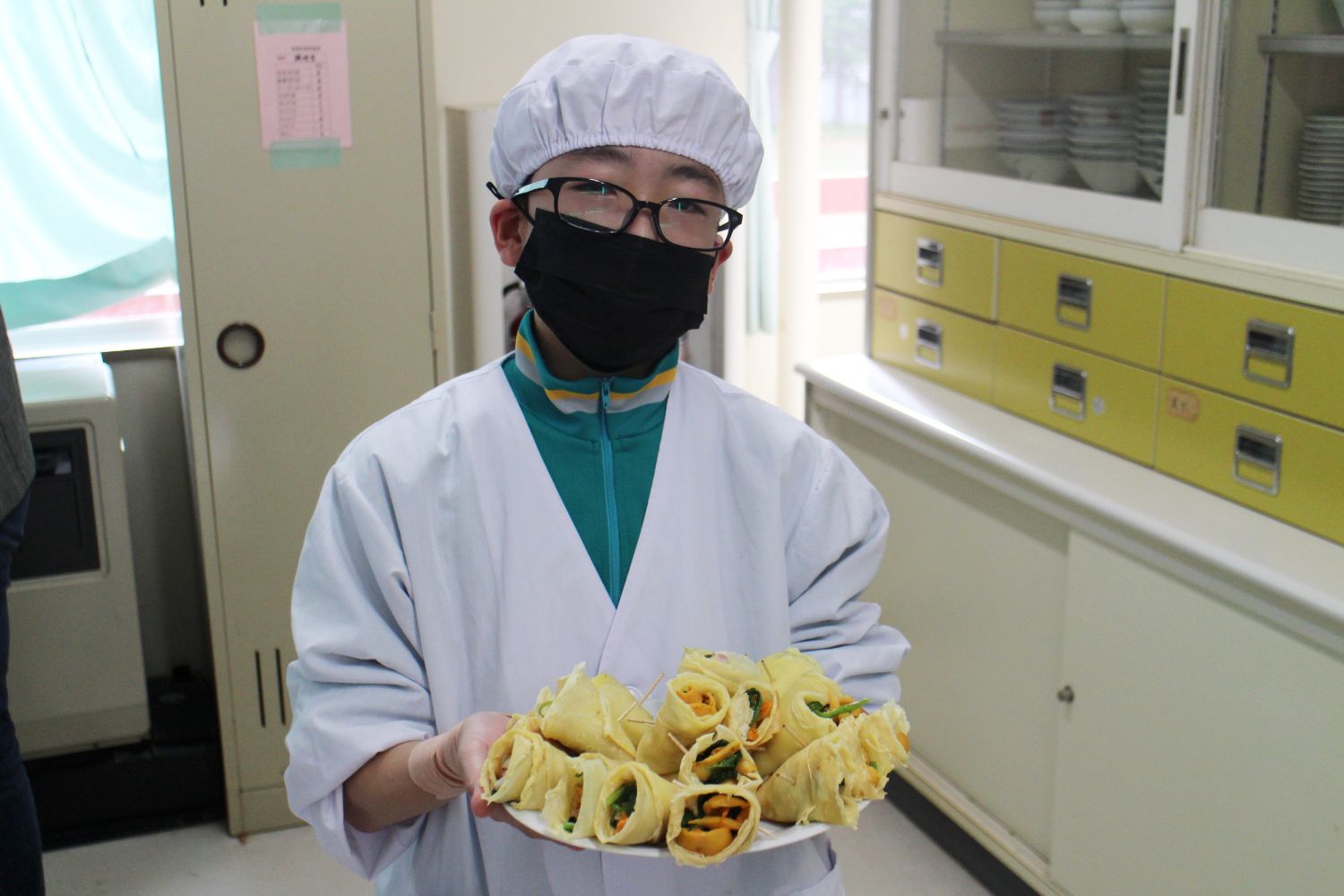 白衣と帽子をかぶった男子生徒が黒いマスクをつけて、完成した具材を巻いたクレープを手に持ちカメラに向かって見せている写真
