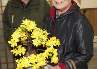 黄色のツツジの繭細工を両手で持ち笑顔の女性の写真