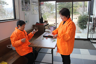 入口のすぐ内側にある備え付けの木のベンチに座った女性と、長机越しに向かい合って立った女性が、おそろいのオレンジ色のジャンバーを着て、絹糸を巻く作業をしている写真