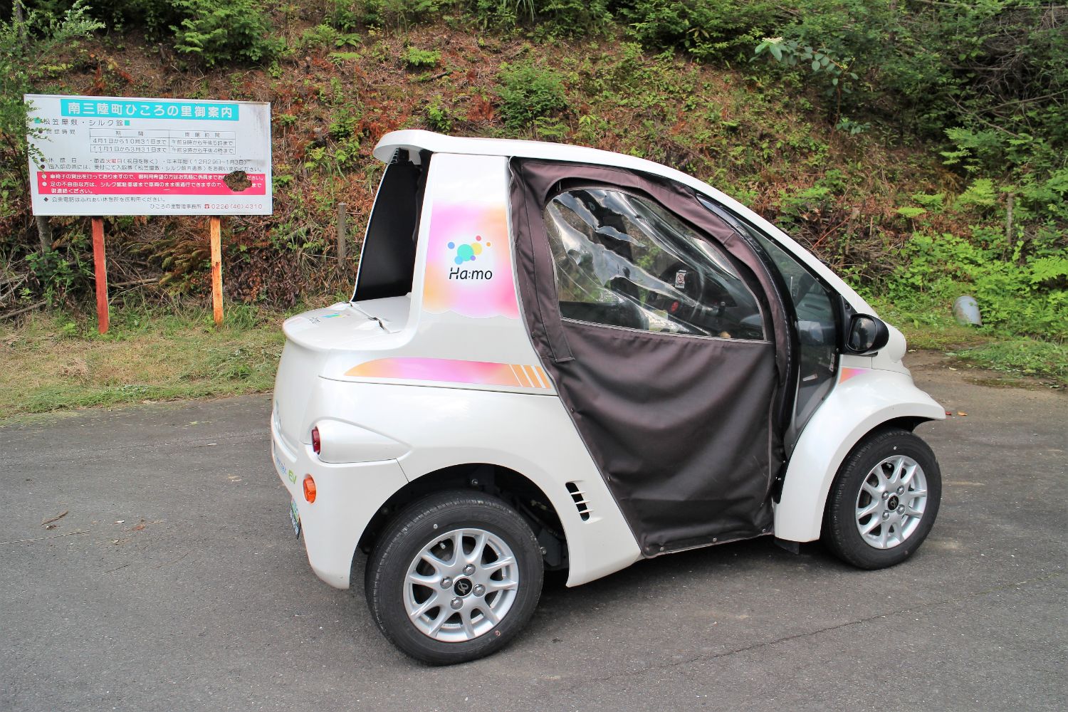 山あいの案内看板と緑に囲まれた駐車場に、ドアカバーが閉められた白い一人乗り超小型EVが停まっている写真